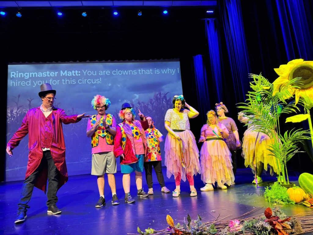 performers dressed as clowns and a ringmaster on stage with a large screen behind with lines displayed