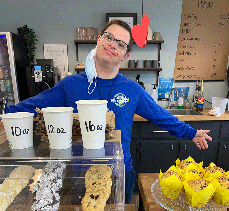 bakery team member wearing a blue team Prescott shirt smiles while standing behind the bakery counter with their arms wide open in a welcoming gesture
