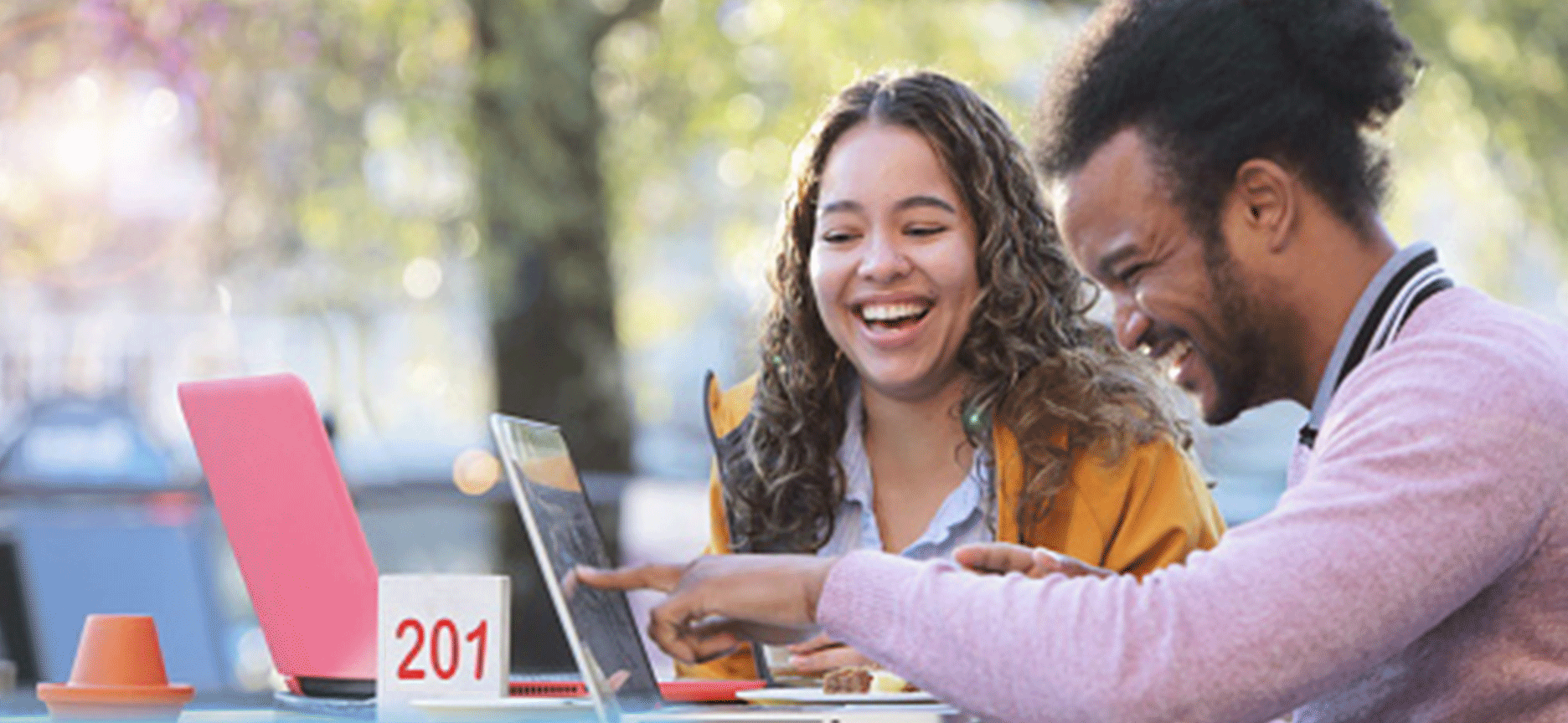 two people sitting together at an outdoor cafe table with laptops, sharing conversation.