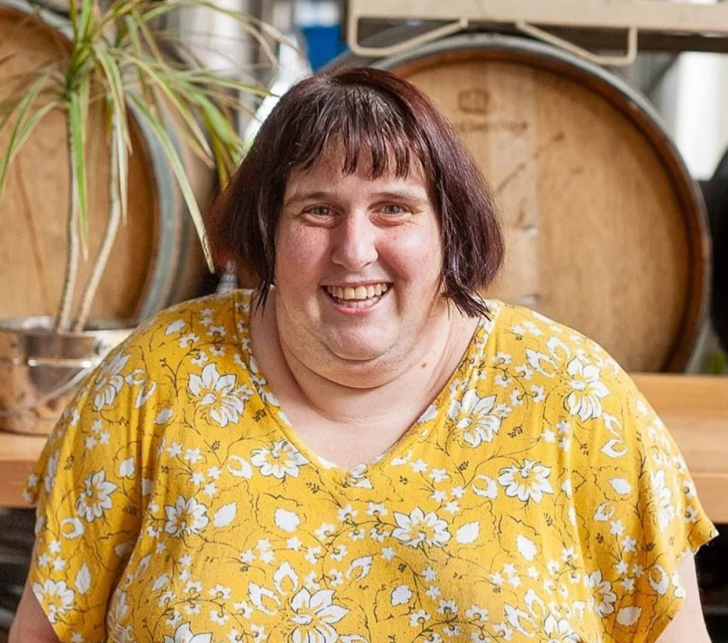 individual smiles while standing in front wooden barrels and a long table.